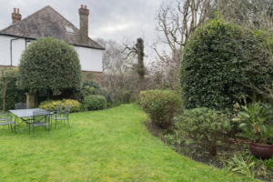 Neat backyard garden with green lawn, patio table and chairs, and mature shrubs beside a white house on a cloudy day