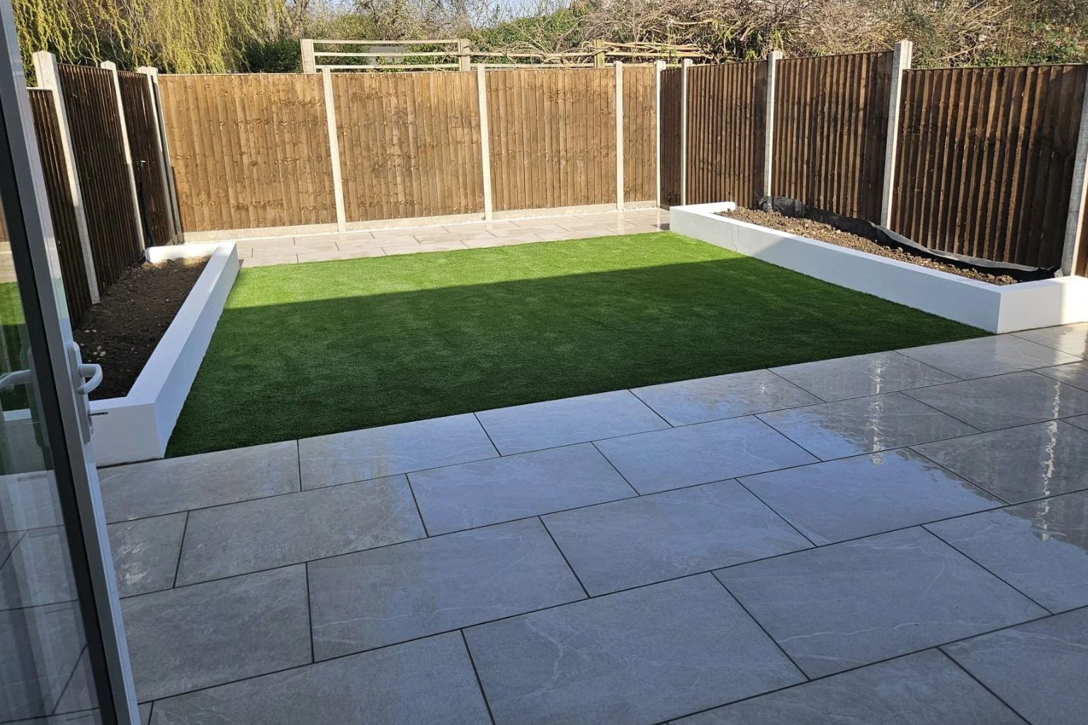 After photo of a fully landscaped back garden featuring a lush artificial grass lawn, large light grey porcelain patio slabs, white rendered raised borders, and a bamboo panel fence with trees visible above