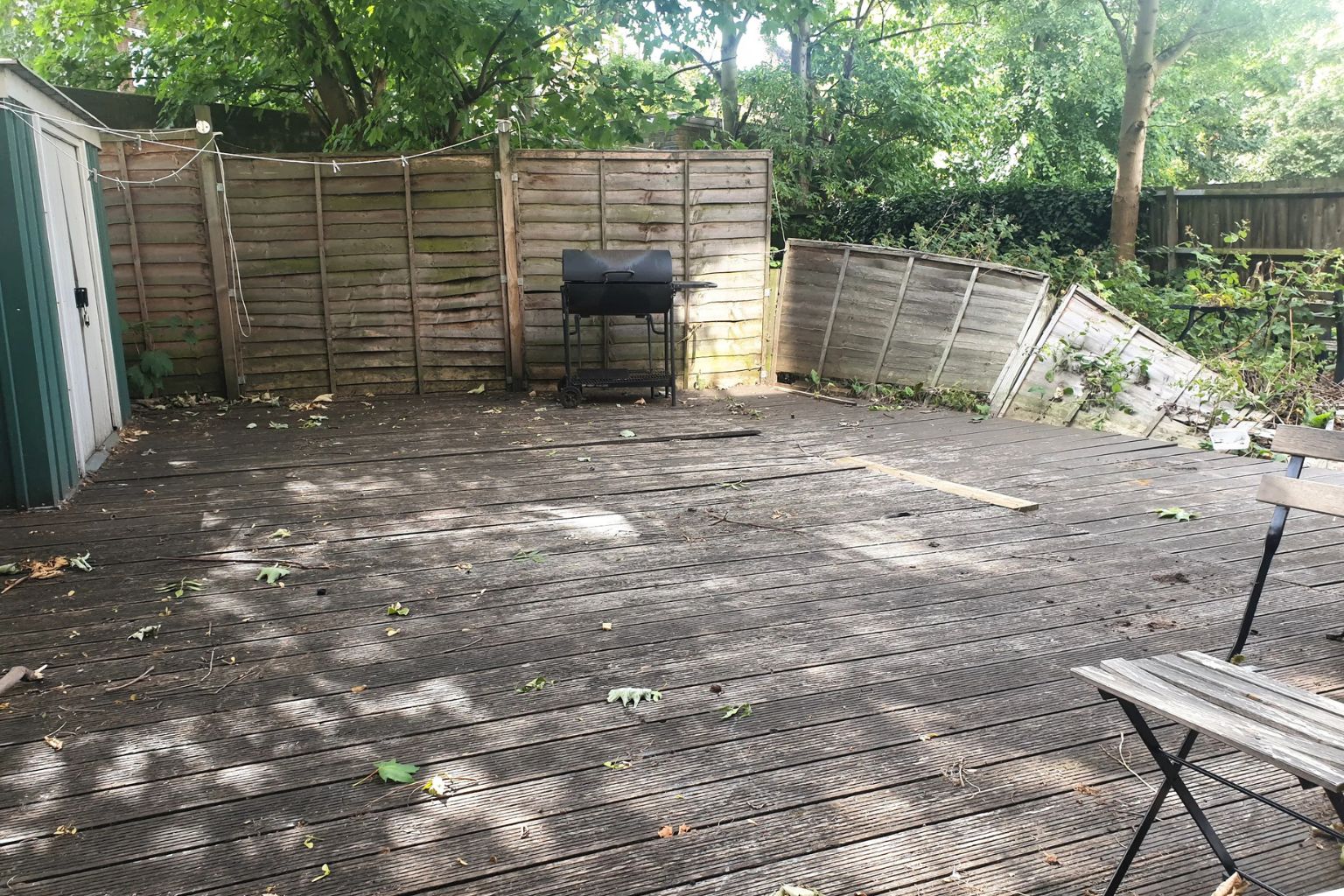 Neglected back garden in South East London with worn and weathered wooden decking, an old wooden panel fence, overgrown vegetation along the borders, and a black barbecue grill in the corner.