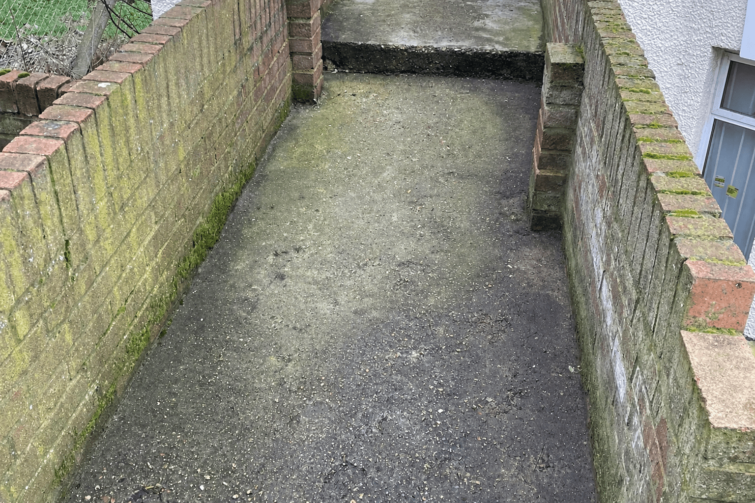 Before photo of a narrow concrete pathway between two moss-covered brick walls in South East London, prior to jet washing service by Urban Gardeners.