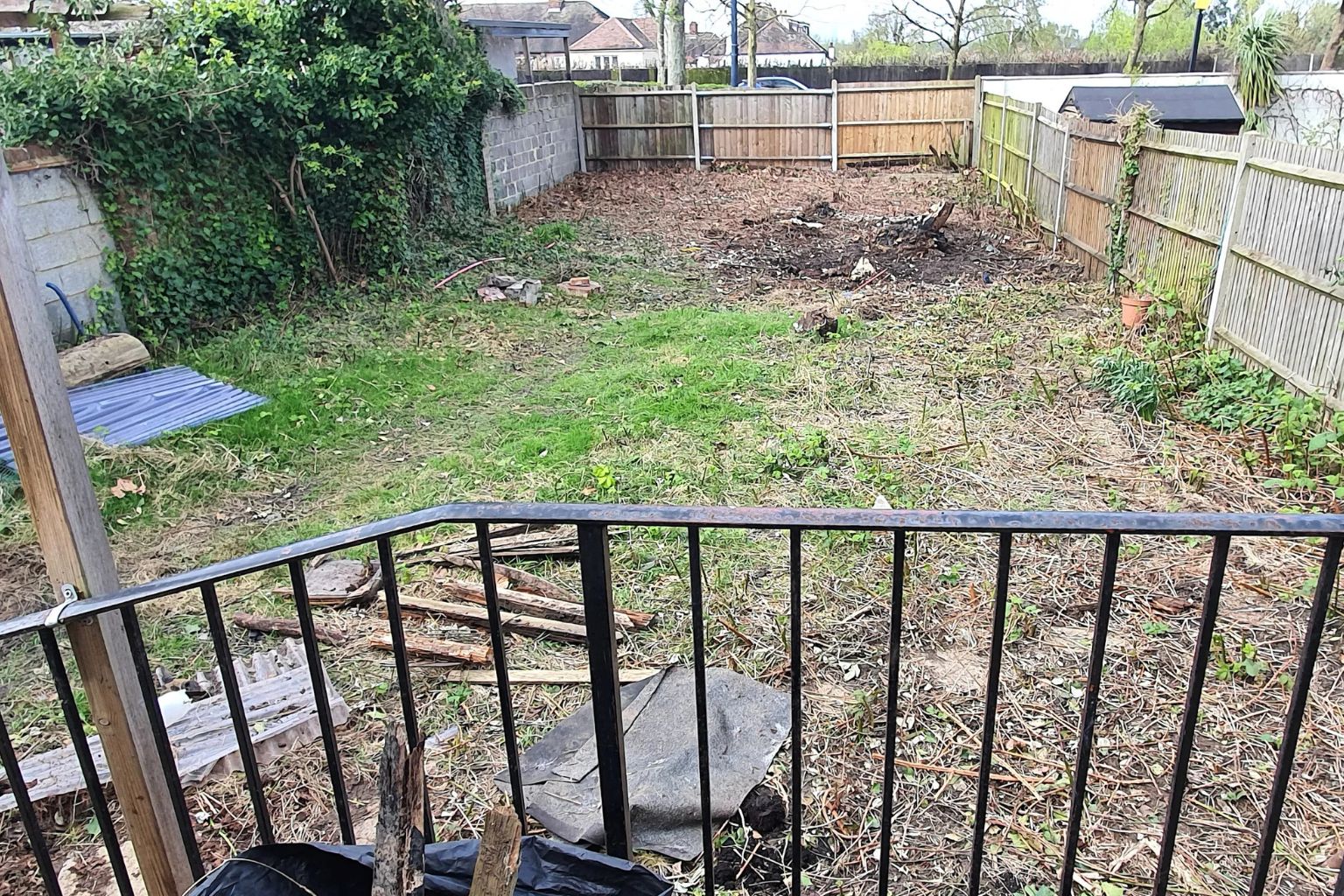 efore photo of an overgrown and neglected back garden with patchy grass, bare soil, and a metal railing in the foreground