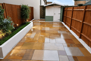 Modern renovated backyard patio with large glossy stone tiles, wooden privacy fencing, raised white planter beds with greenery, soft landscape lighting, and a small garden shed in the background on a bright clean day.